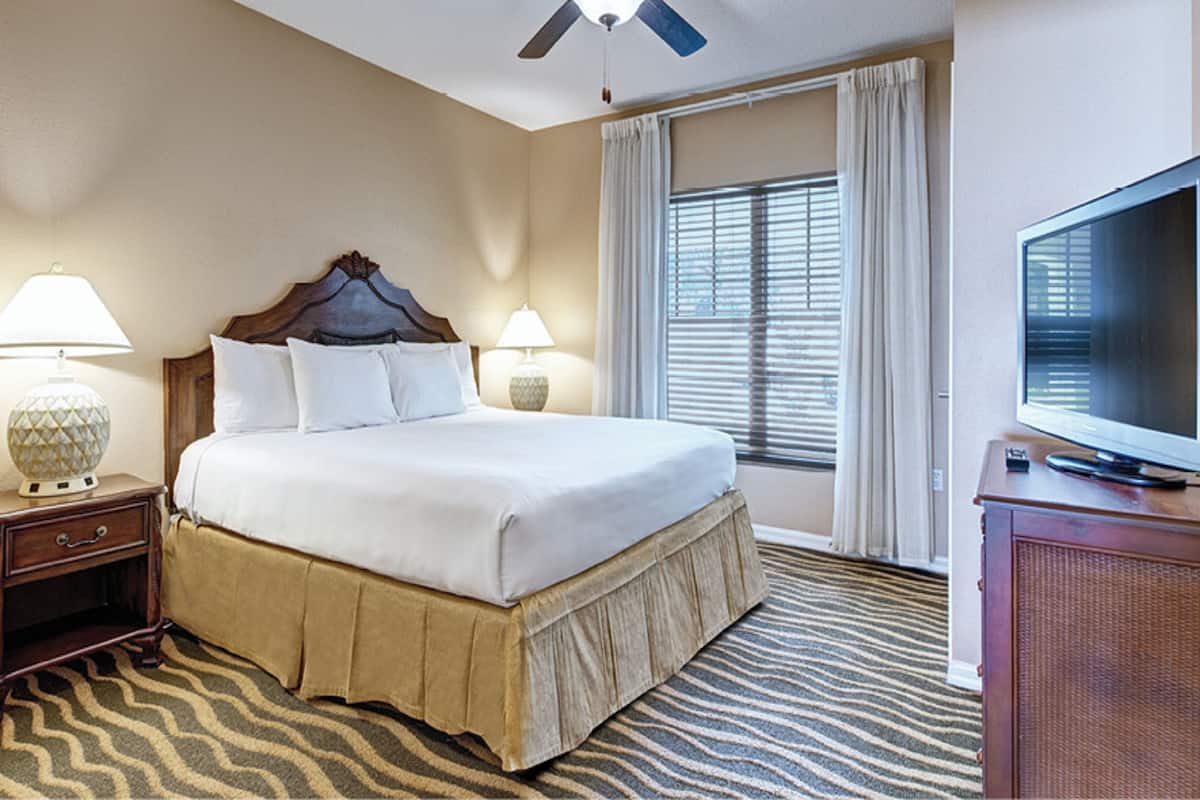 Una habitación de hotel con una cama grande, dos lámparas de noche, un televisor sobre un mueble de madera y una ventana con cortinas.