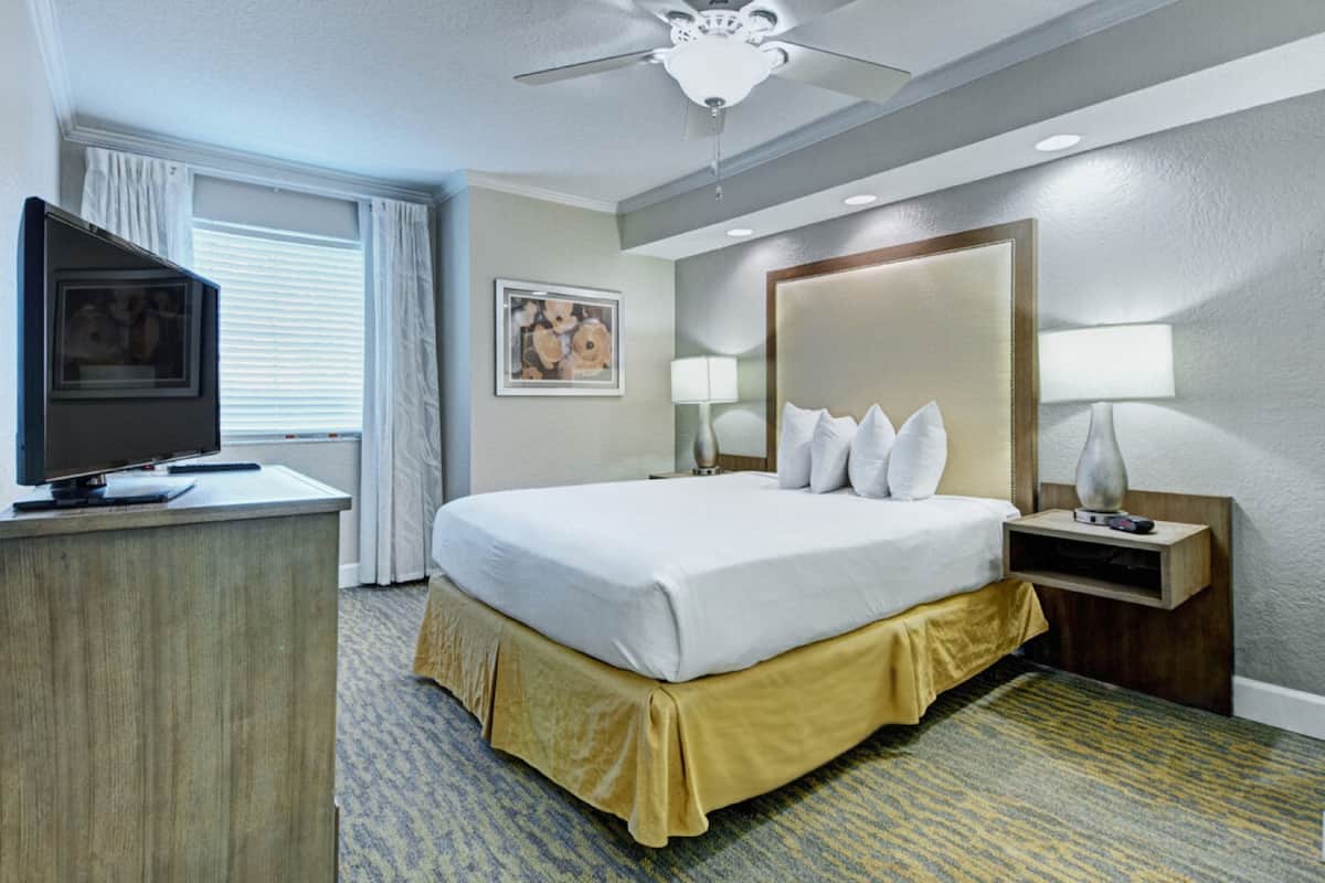 Una habitación de hotel con cama, escritorio con televisor, ventilador de techo, mesita de noche con lámpara y un cuadro enmarcado en la pared.