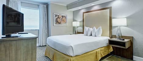 Quarto de hotel com cama, escrivaninha com televisão, ventilador de teto, mesa de cabeceira com abajur e quadro na parede.
