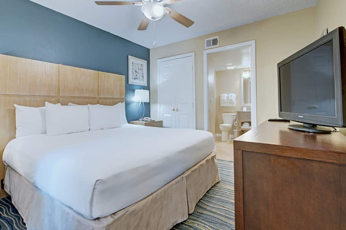 Habitación de hotel con una cama grande, cabecera de madera, televisor sobre un aparador de madera y baño visible a través de una puerta abierta.
