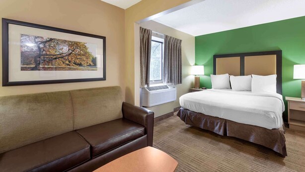 A hotel room with a bed, a couch, a window with curtains, an air conditioning unit, and a framed picture on the wall. - Extended Stay America Suites Cleveland Westlake (Westlake)