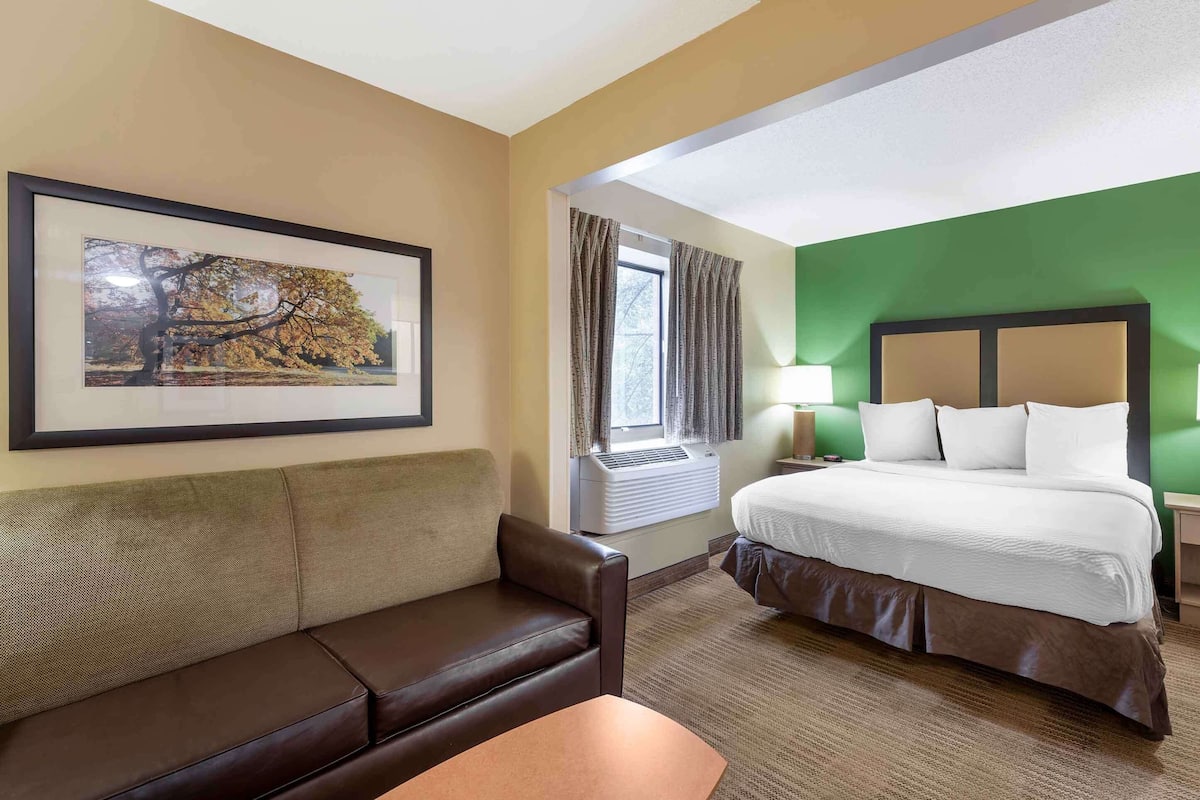 Una camera d'albergo con un letto, un divano, una finestra con tende, un condizionatore d'aria e un quadro appeso al muro.