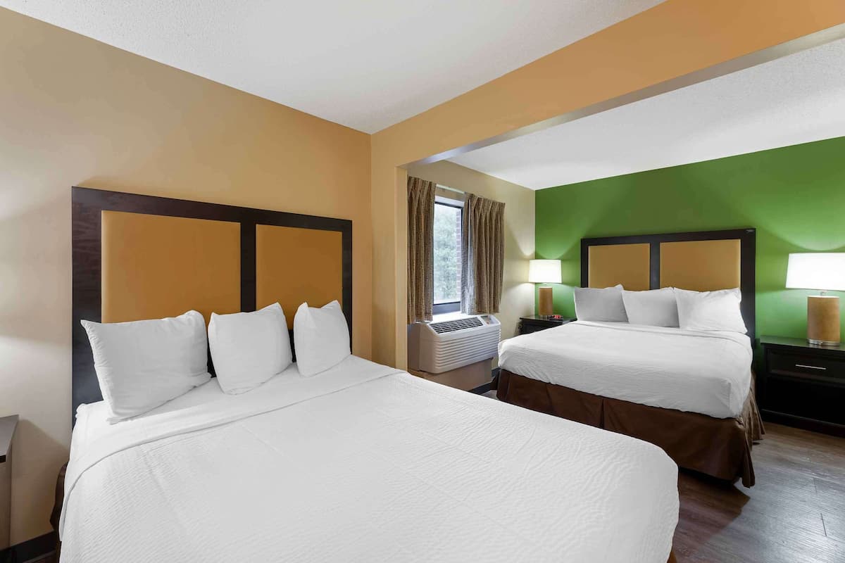 Una camera d'albergo con due letti, una finestra con tende, un condizionatore d'aria e un comodino con una lampada.