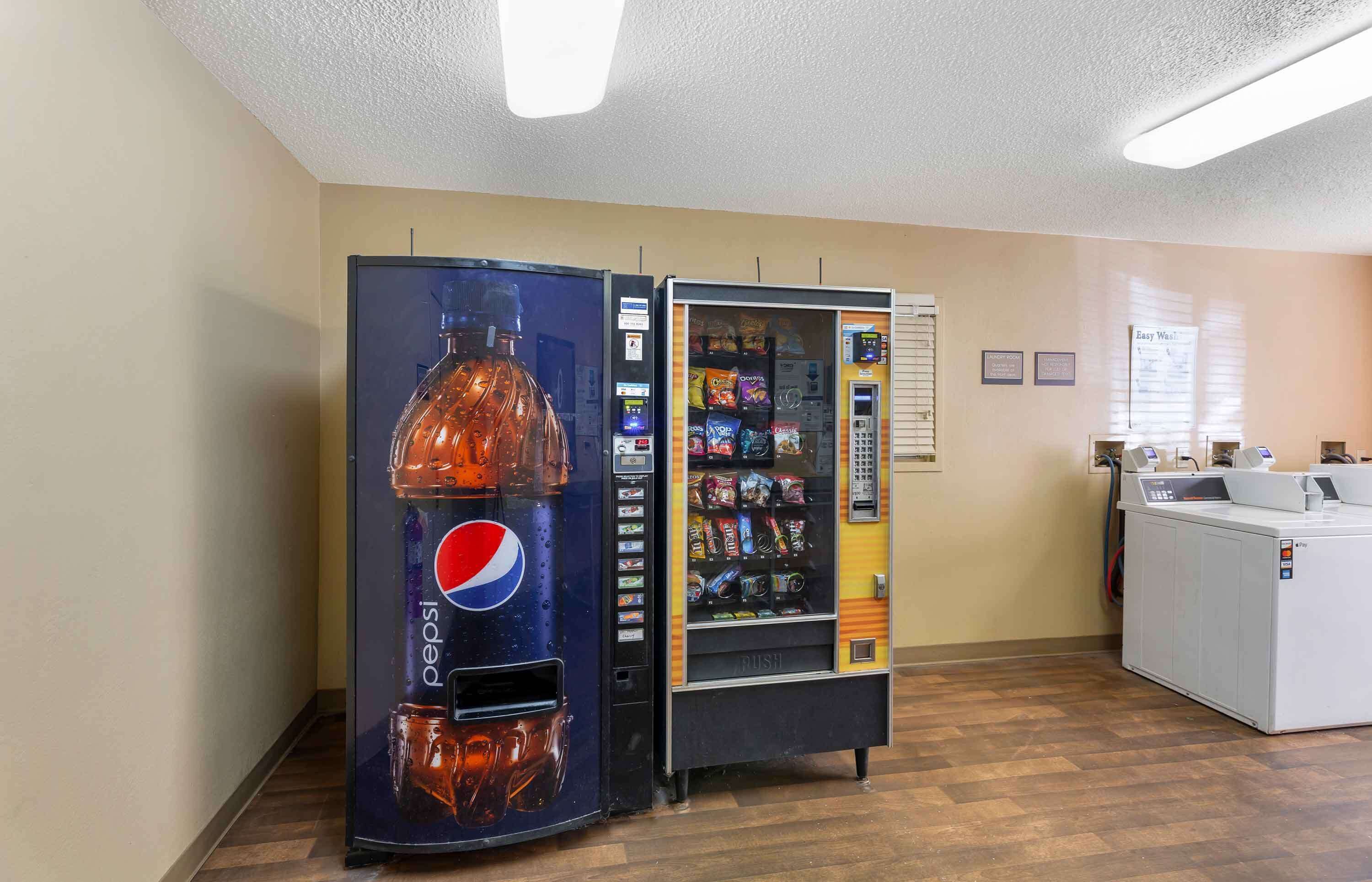 A vending machine with a Pepsi logo, a microwave, and a washing machine in a room with wooden flooring and beige walls.