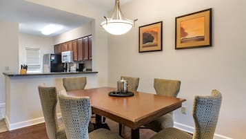 A dining area with a wooden table and six patterned chairs. There is a kitchen in the background with wooden cabinets and a microwave. Two framed pictures are on the wall above the table.