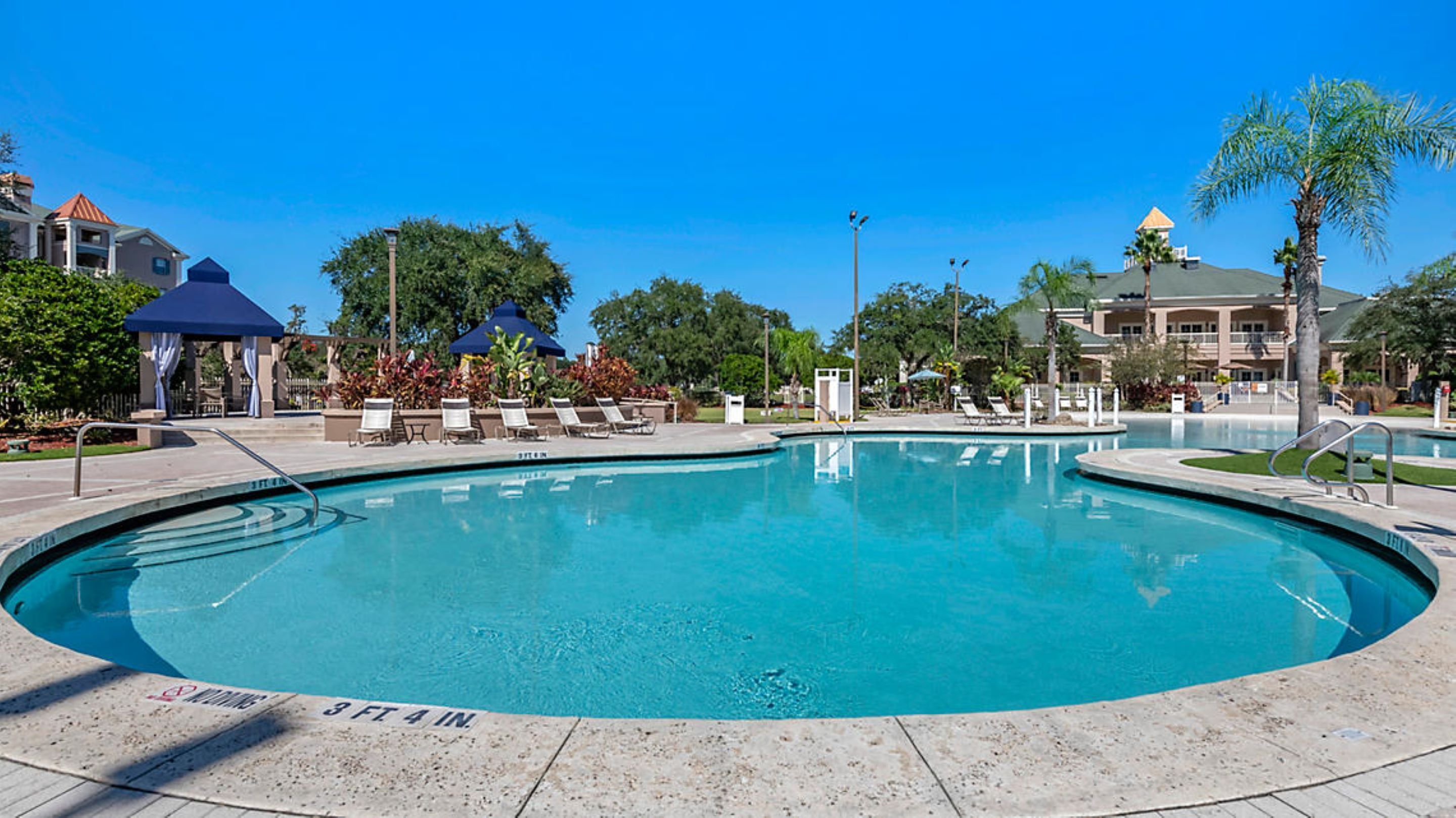 A large, clear swimming pool with lounge chairs and umbrellas, surrounded by palm trees and buildings.
