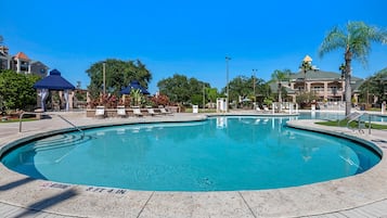 A large, clear swimming pool with lounge chairs and umbrellas, surrounded by palm trees and buildings.