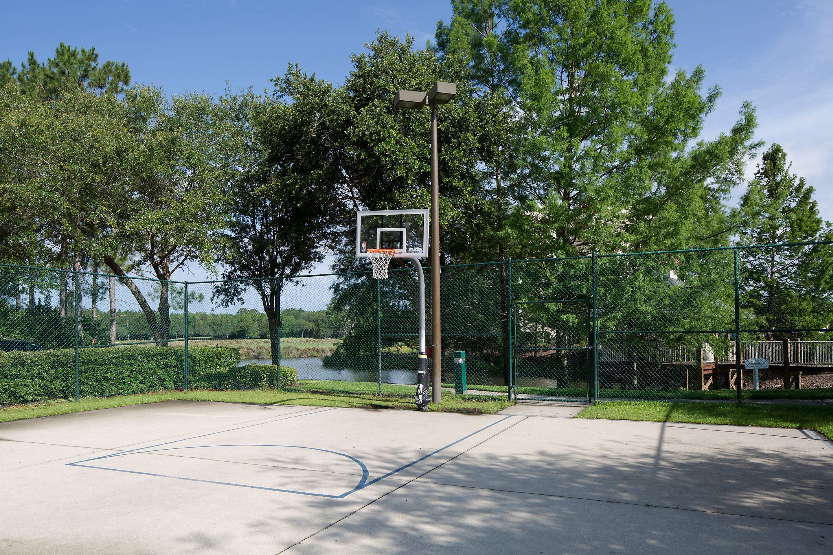 An outdoor basketball court with a hoop, surrounded by a fence and trees.