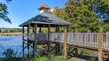 A wooden gazebo with a tiled roof, situated on stilts over a body of water, surrounded by trees and vegetation.