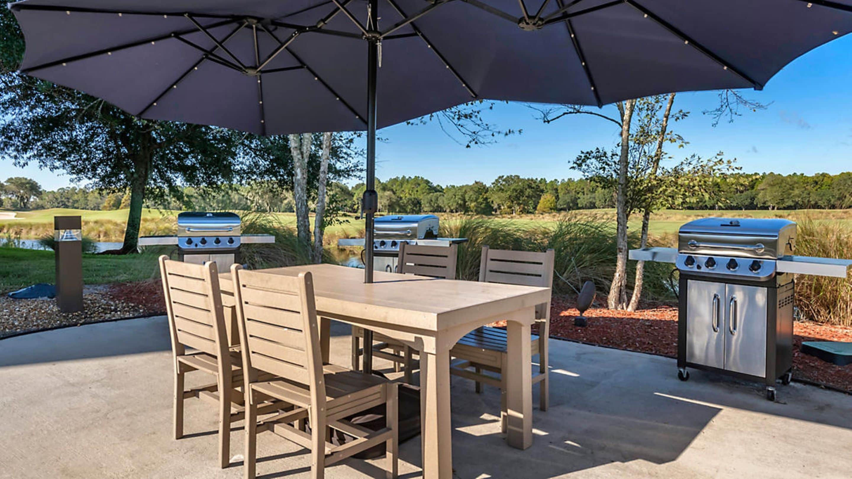 An outdoor patio with a table and chairs, two barbecue grills, and a large umbrella providing shade.