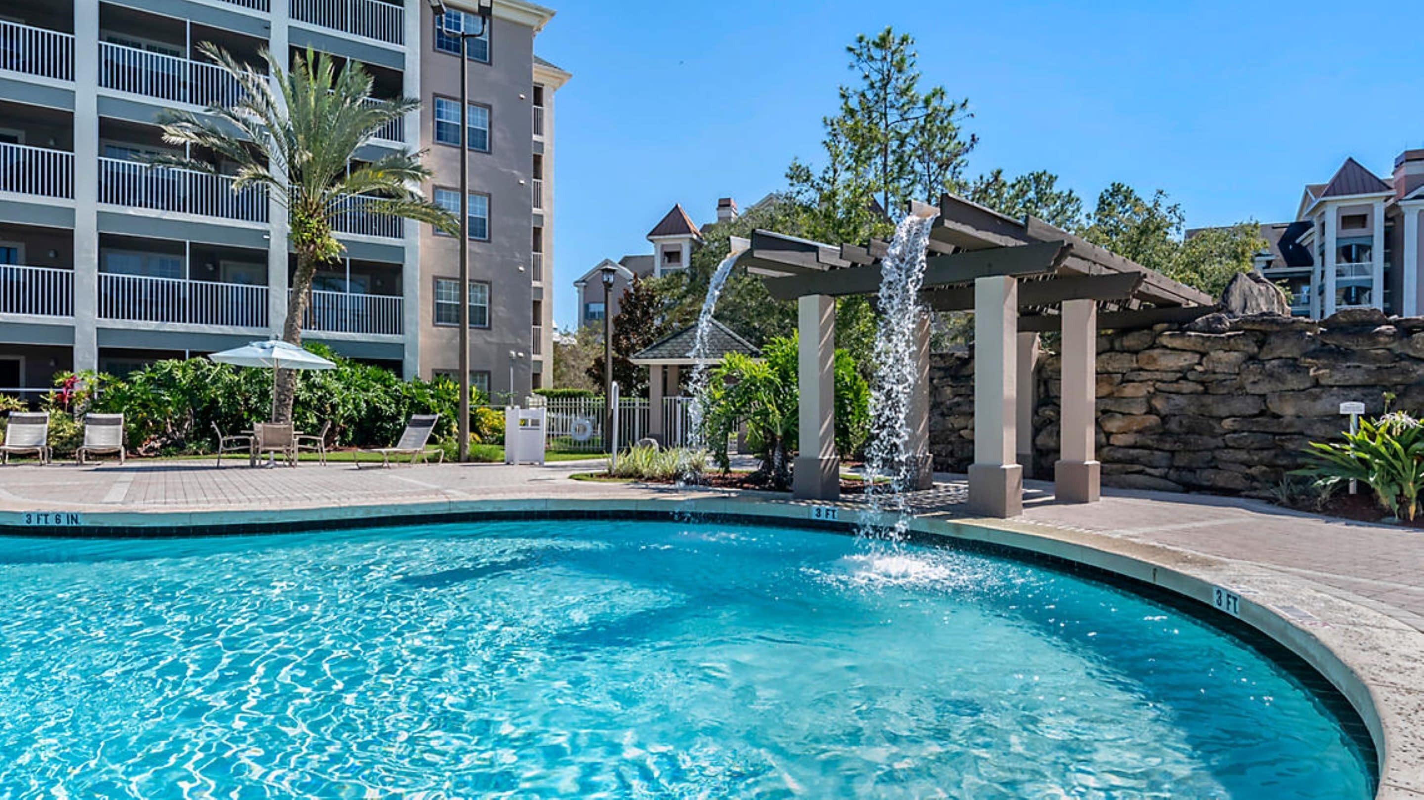 A swimming pool with a waterfall feature, surrounded by a stone wall and a multi-story building with balconies.