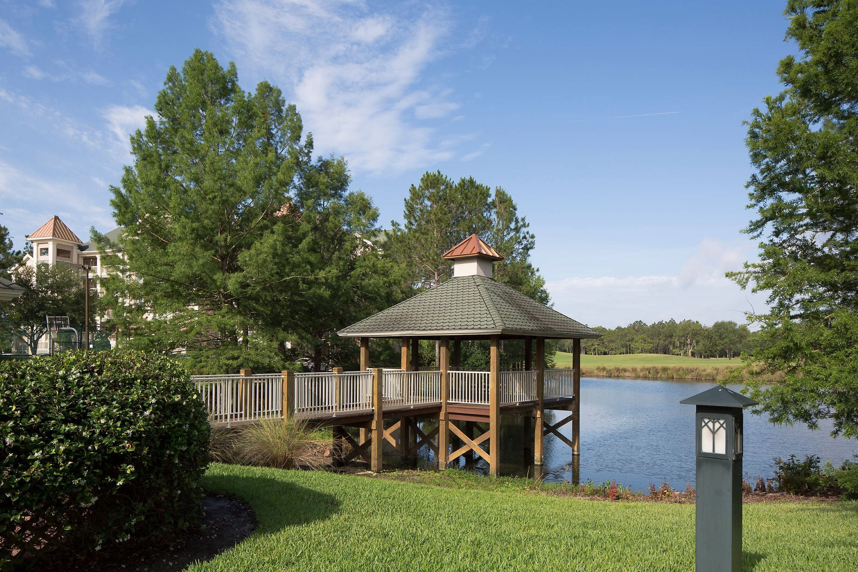 A gazebo with a wooden deck over a body of water, surrounded by trees and a well-maintained lawn.