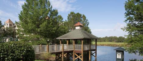 A gazebo with a wooden deck over a body of water, surrounded by trees and a well-maintained lawn.