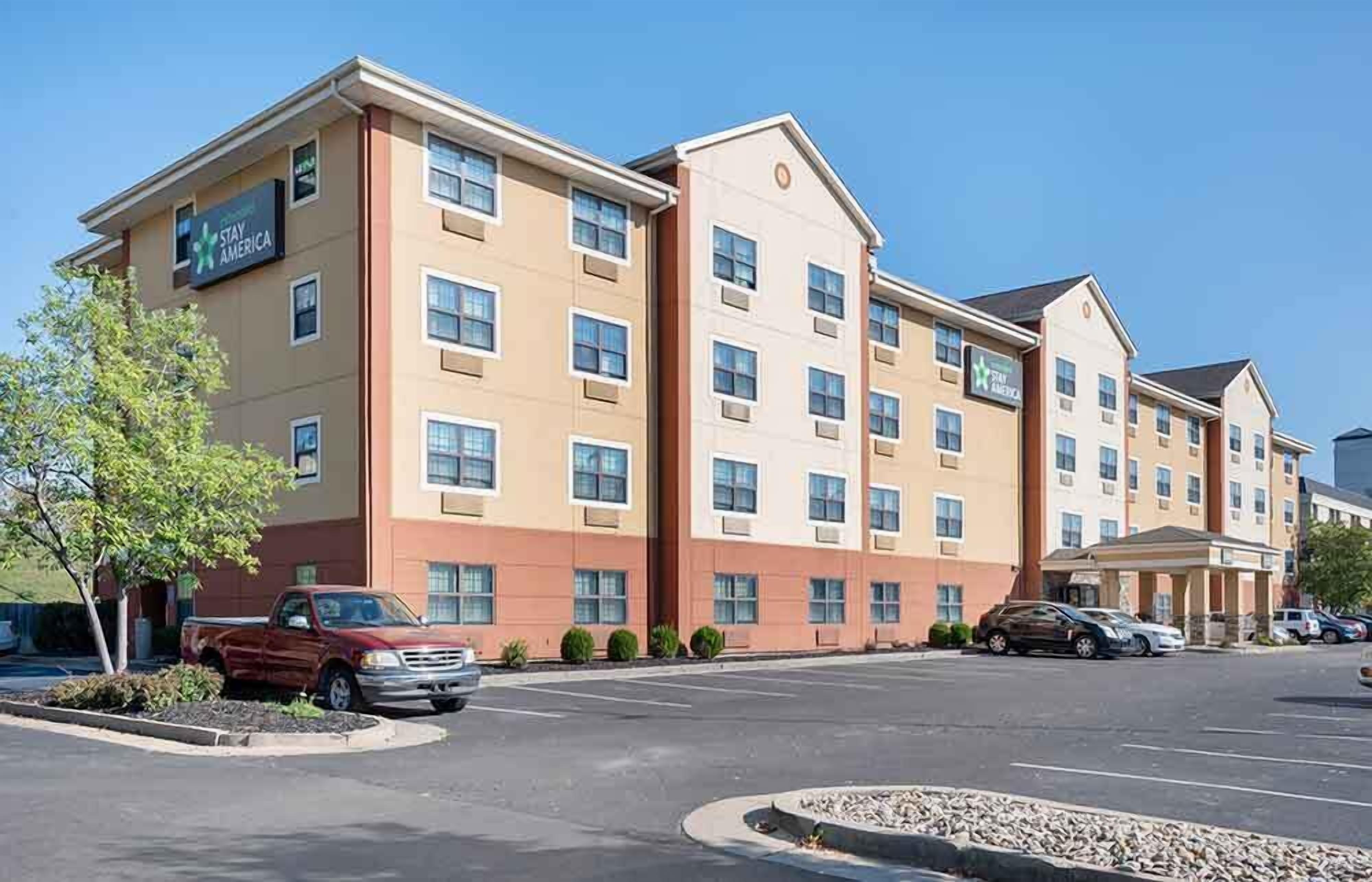 A Stay America hotel with a multi-story building, parking lot, and a few cars parked in front.