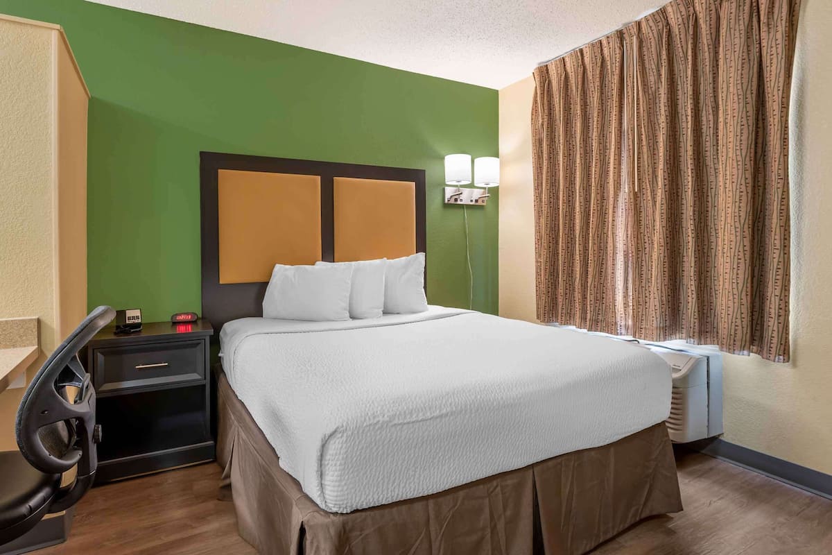 A hotel room with a bed, a nightstand, a chair, and a window with curtains.
