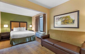 Habitación de hotel con cama, sofá, ventana con cortinas, unidad de aire acondicionado y un cuadro en la pared.