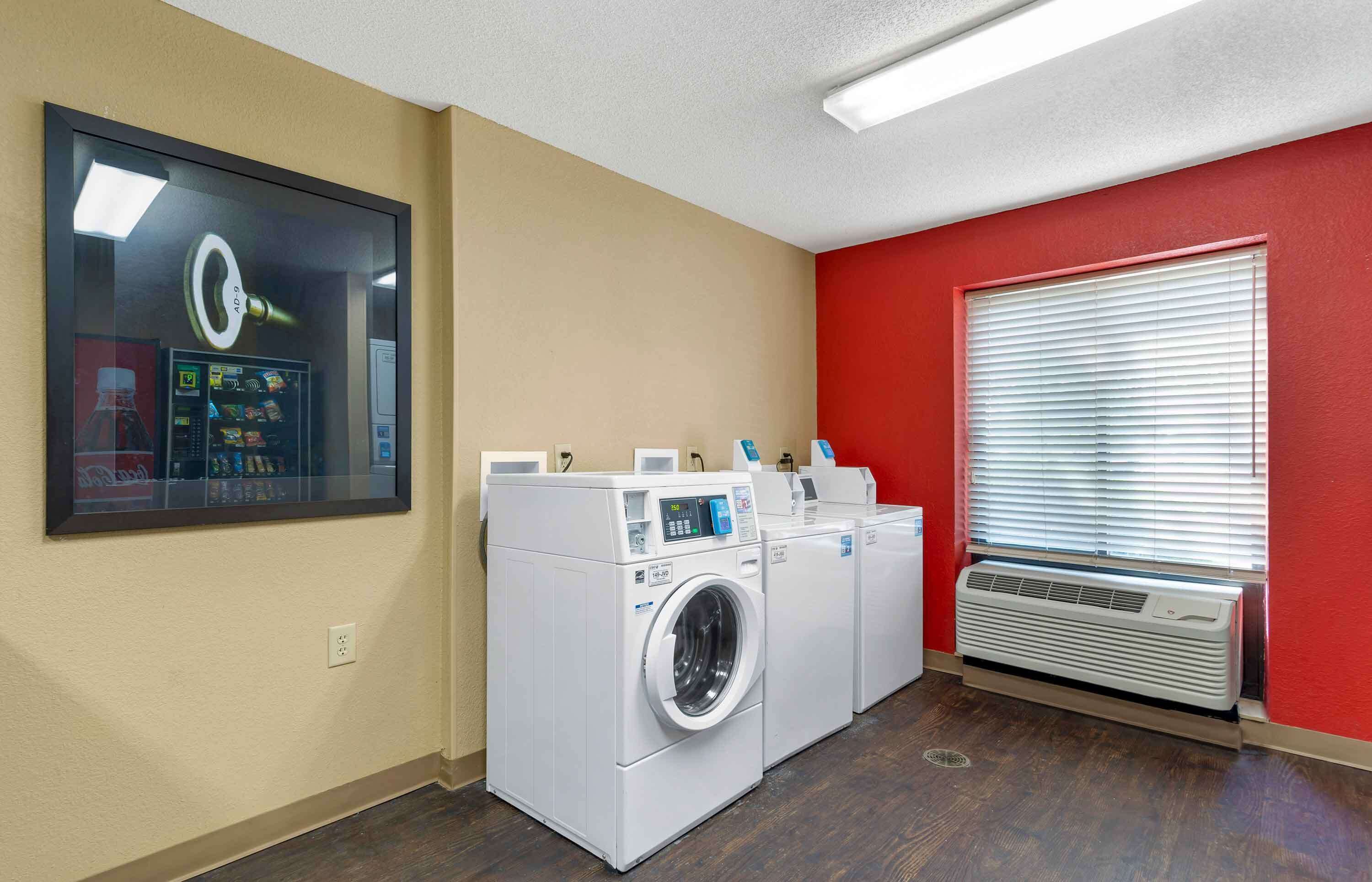 Une buanderie équipée de deux machines à laver à chargement frontal, d’un sèche-linge fixé au mur et d’une grande fenêtre avec des stores.
