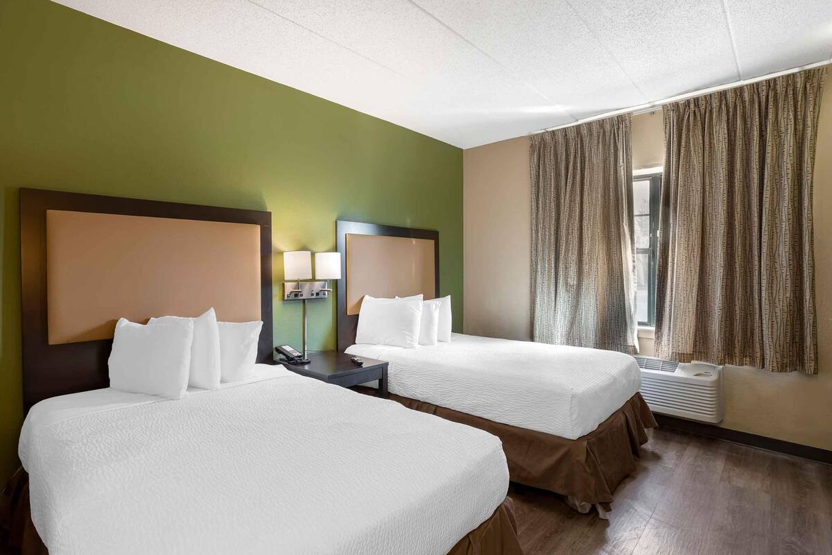 A hotel room with two beds, a nightstand, a lamp, and a window with curtains.