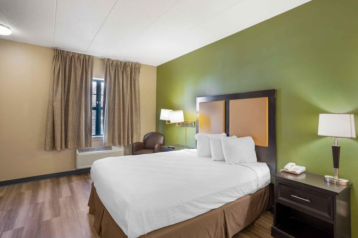 A hotel room with a large bed, a desk with a lamp, a chair, and a window with curtains.