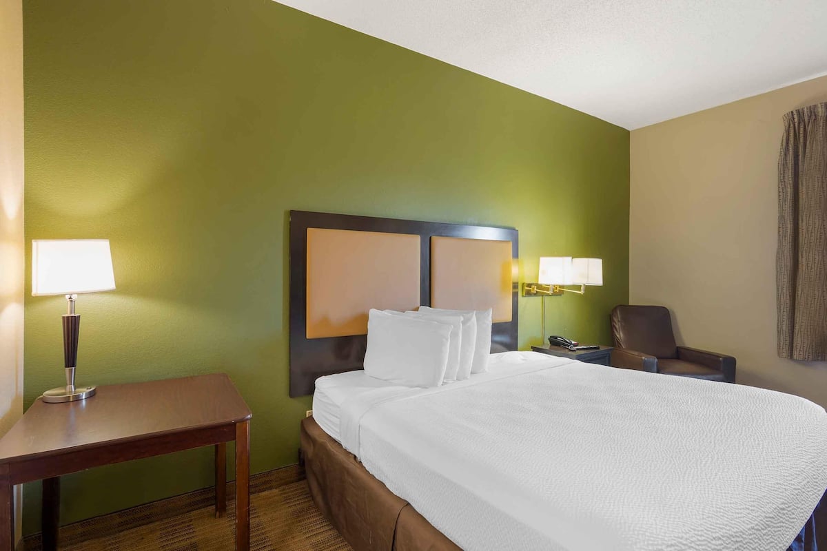 A hotel room with a large bed, two bedside lamps, a small table, a chair, and a headboard with a light fixture.