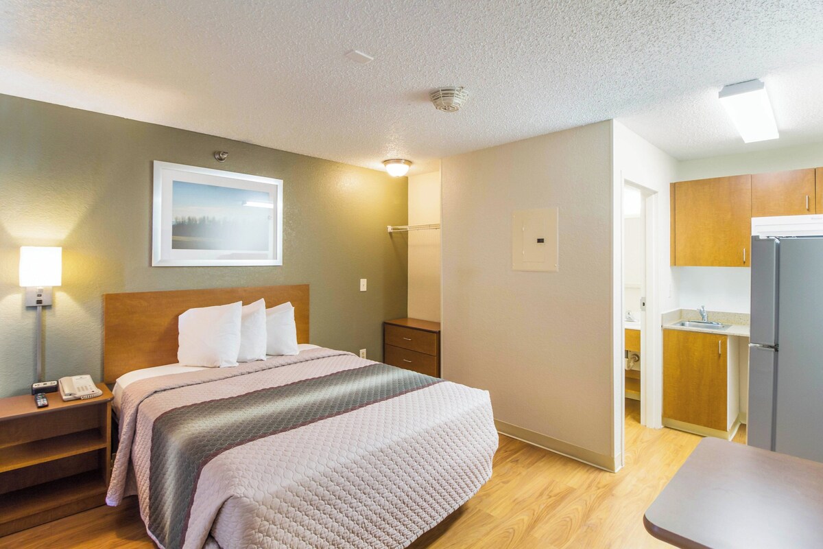 Habitación de hotel con cama, mesita de noche, lámpara y vista al área de la cocina.