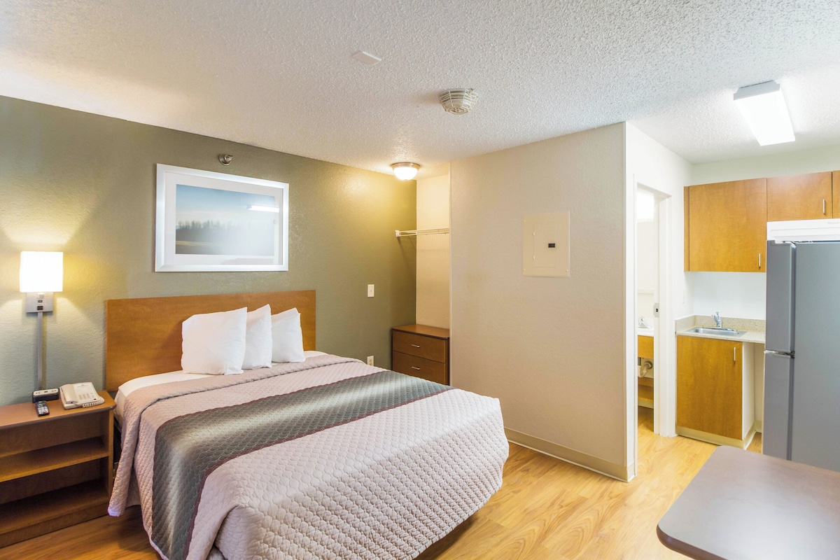 A hotel room with a bed, bedside table, lamp, and a view of the kitchen area.