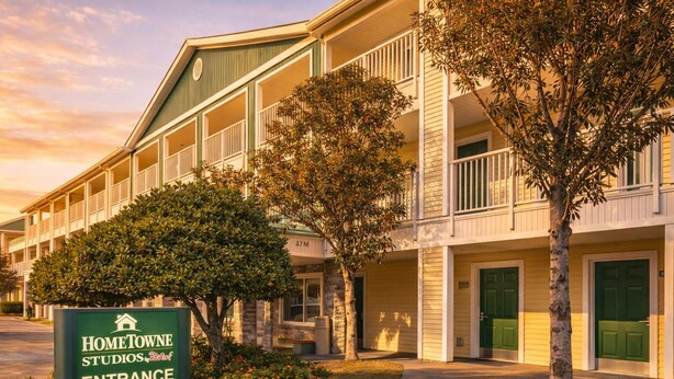 Un hotel de varios pisos con un letrero de entrada verde, balcones y una zona ajardinada con árboles.