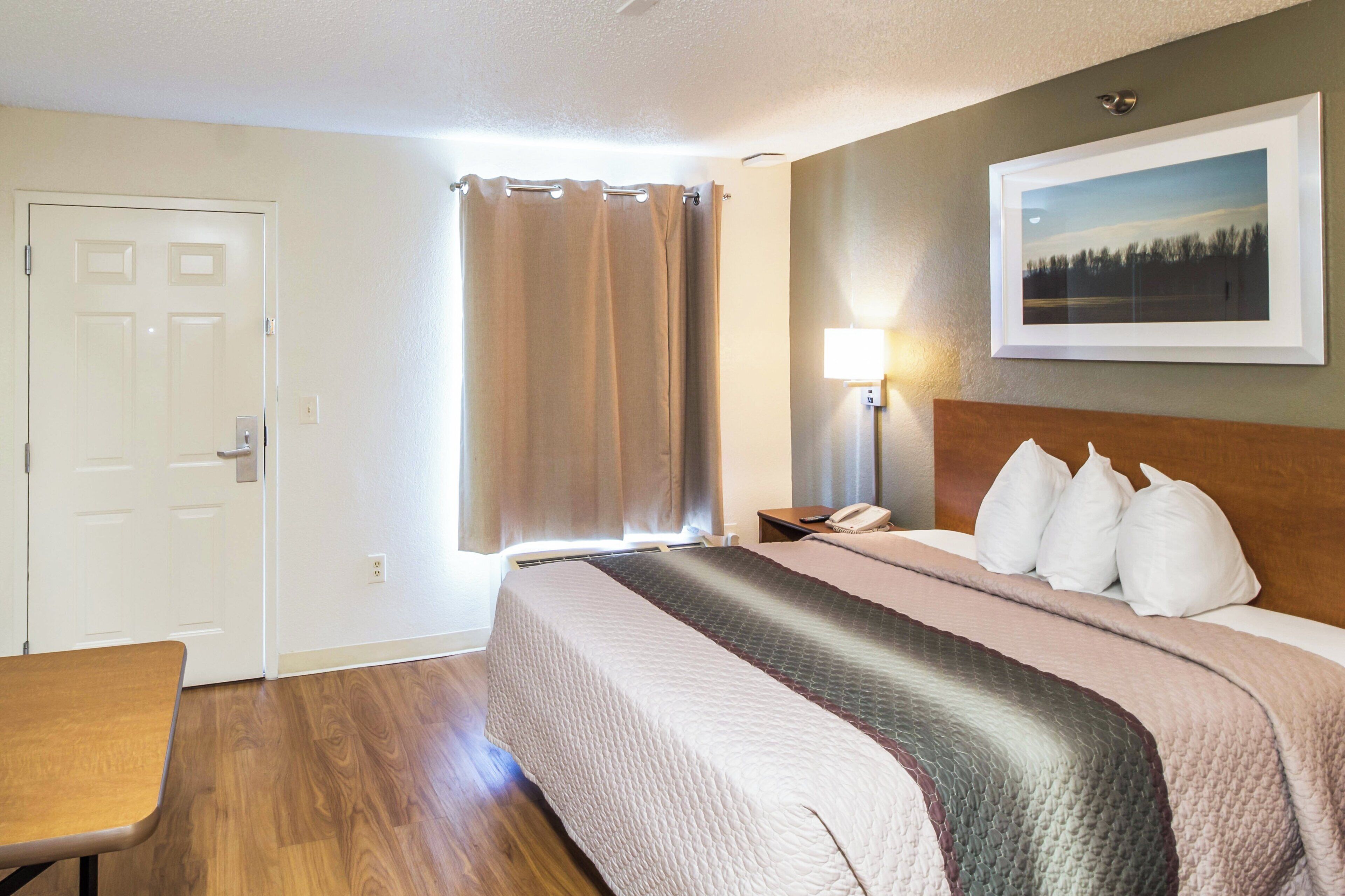 A hotel room with a bed, a desk, a window with curtains, a wall-mounted lamp, and a framed picture.