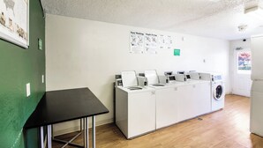 A laundry facility with multiple washing machines and dryers, a table, and informational posters on the wall. - HomeTowne Studios by Red Roof Houston - West Oaks/ Energy Corridor (Houston)