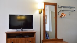 A hotel room with a flat-screen TV on a wooden dresser, a bedside table, a mirror, and a coat rack with hangers. - Value Inn Livonia (Livonia)