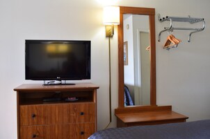 Quarto de hotel com televisão de tela plana sobre uma cômoda de madeira, mesa de cabeceira, espelho e cabideiro com cabides.