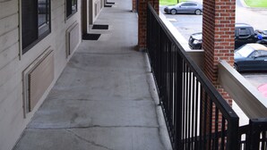 A covered corridor with brick pillars and a black railing, leading to a parking area with cars. - Value Inn Livonia (Livonia)
