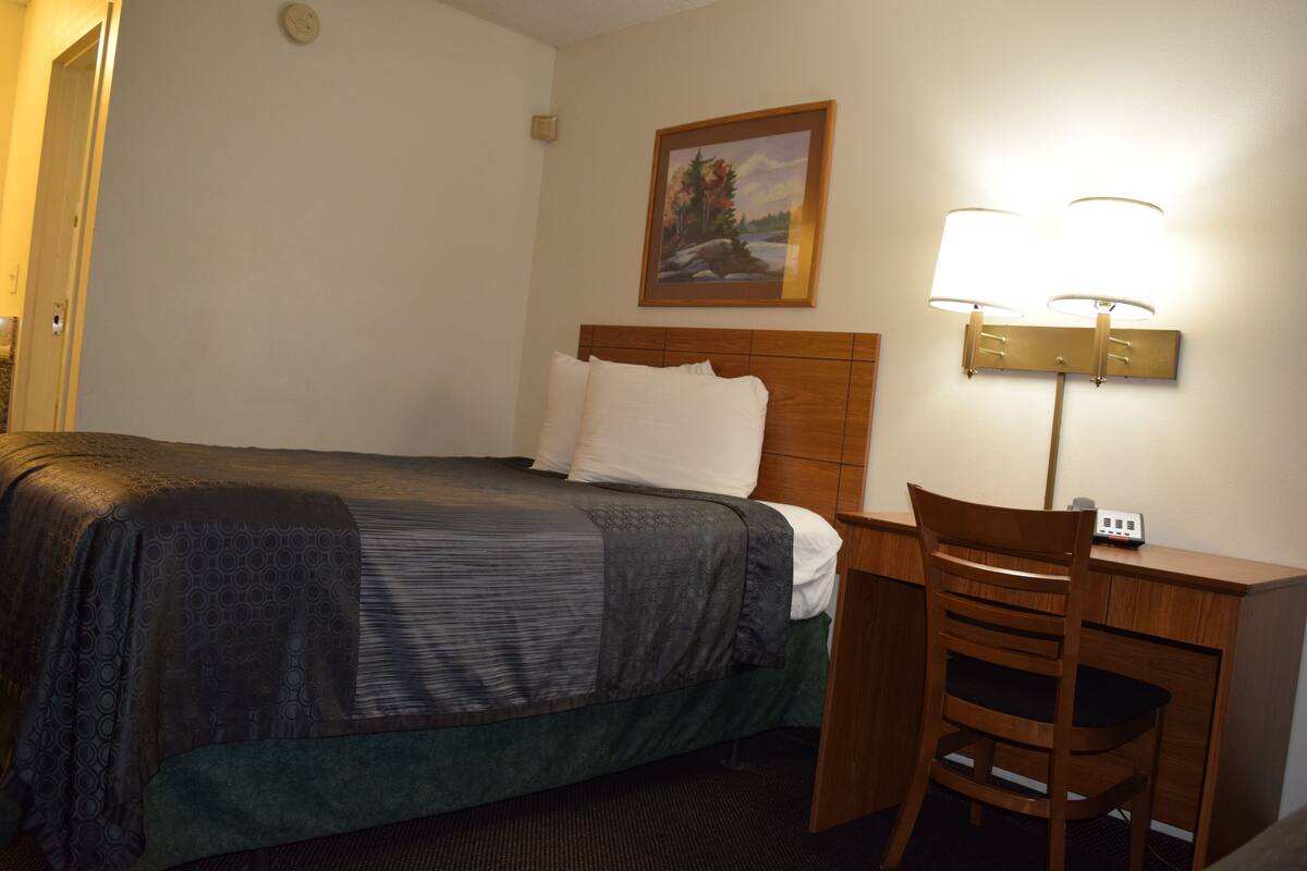 A hotel room with a bed, a desk, a chair, a lamp, and a painting on the wall.