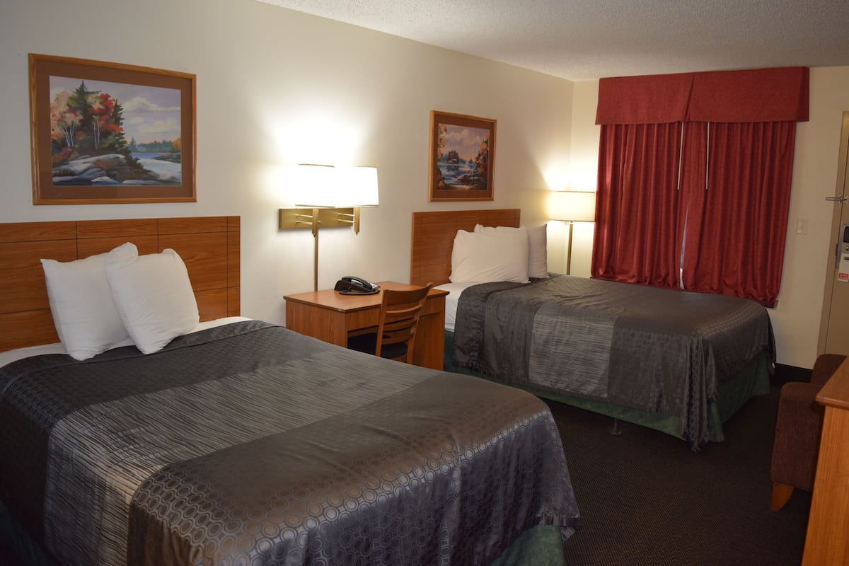 A hotel room with two beds, a desk, a chair, a lamp, and two paintings on the walls.