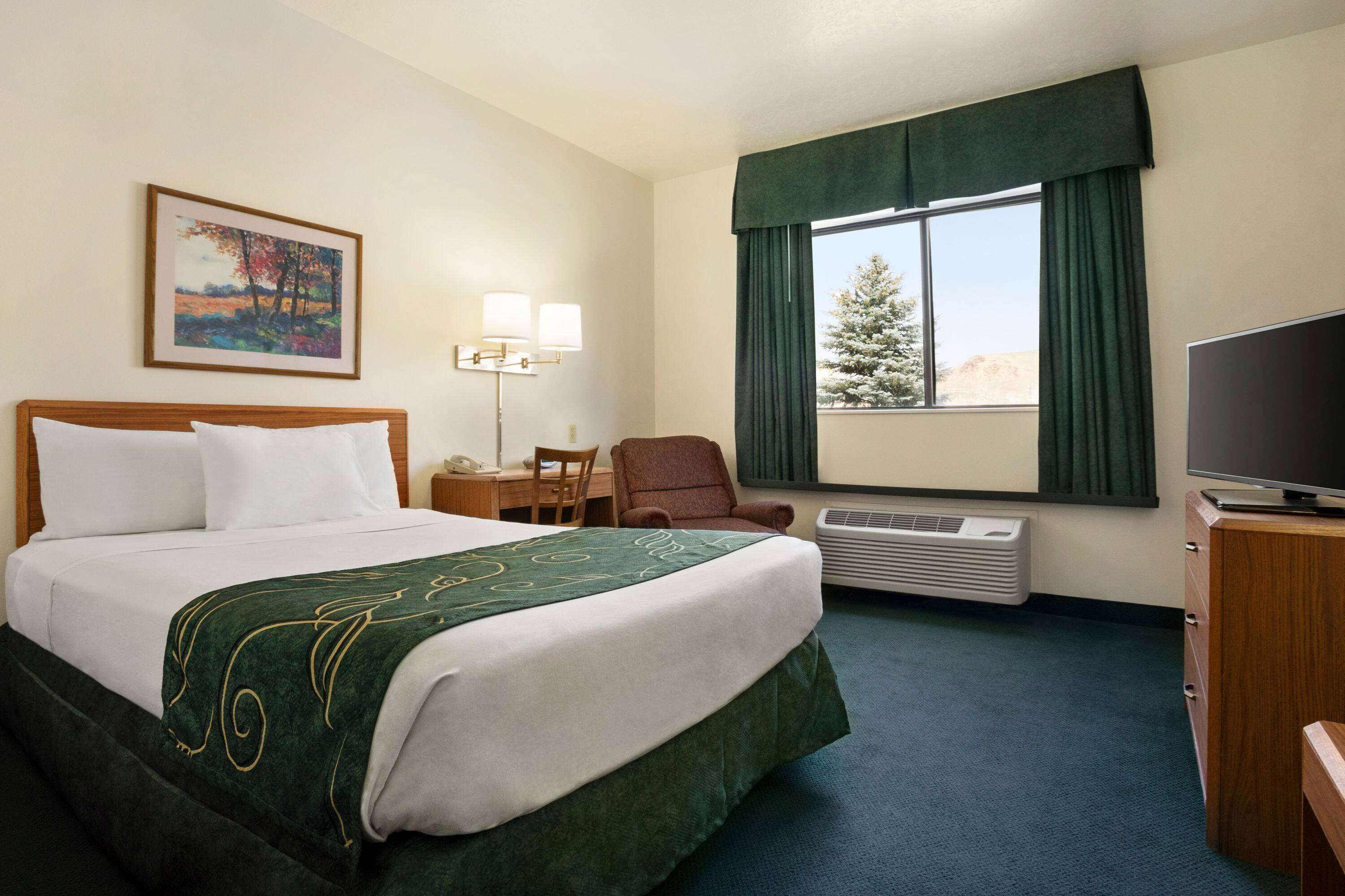 A hotel room with a large bed, a desk with a lamp, a chair, a TV, and a window with curtains.