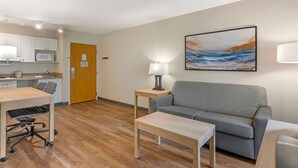 Hotellihuone, jossa on pieni keittiö, harmaa sohva, puinen pöytä, lamppu ja taulu seinällä.