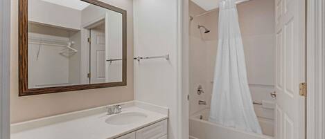 A bathroom with a white vanity, a large mirror, a shower with a white curtain, and a door leading to a walk-in closet.