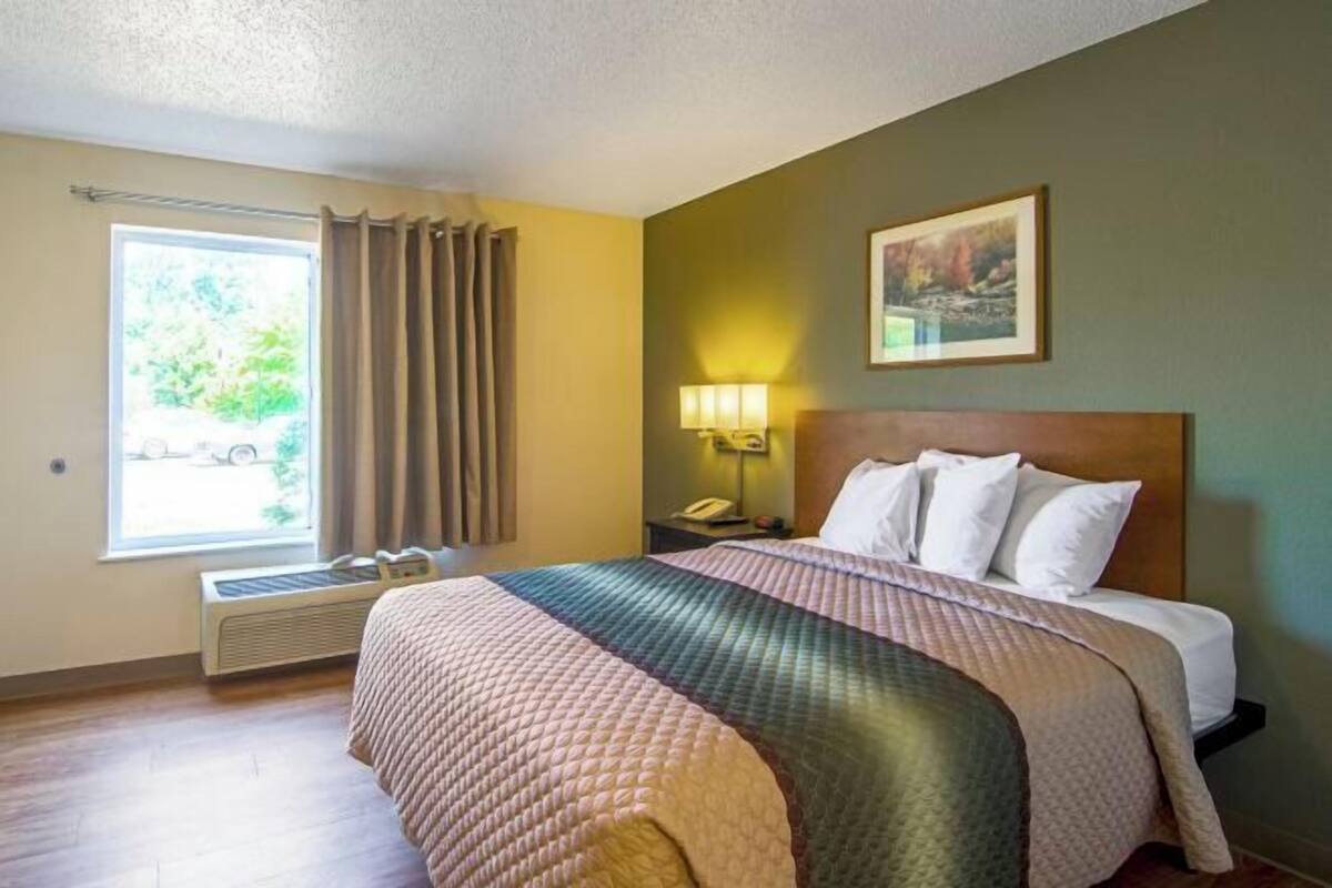 Una habitación de hotel con cama, mesita de noche con lámpara, ventana con cortinas y un cuadro en la pared.