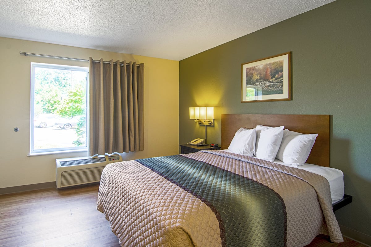 Habitación de hotel con una cama grande, una mesita de noche con lámpara, una ventana con cortinas y un cuadro en la pared.