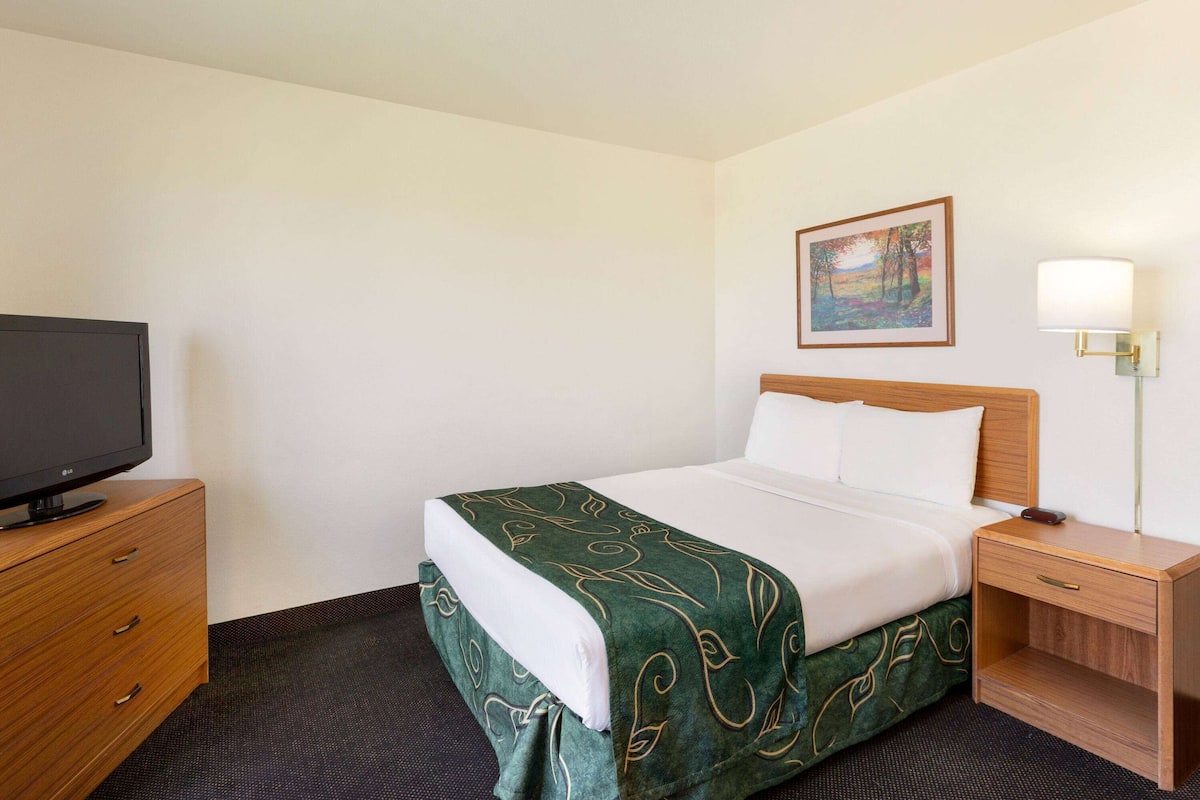 A hotel room with a bed, a TV on a wooden dresser, a nightstand, and a painting on the wall.