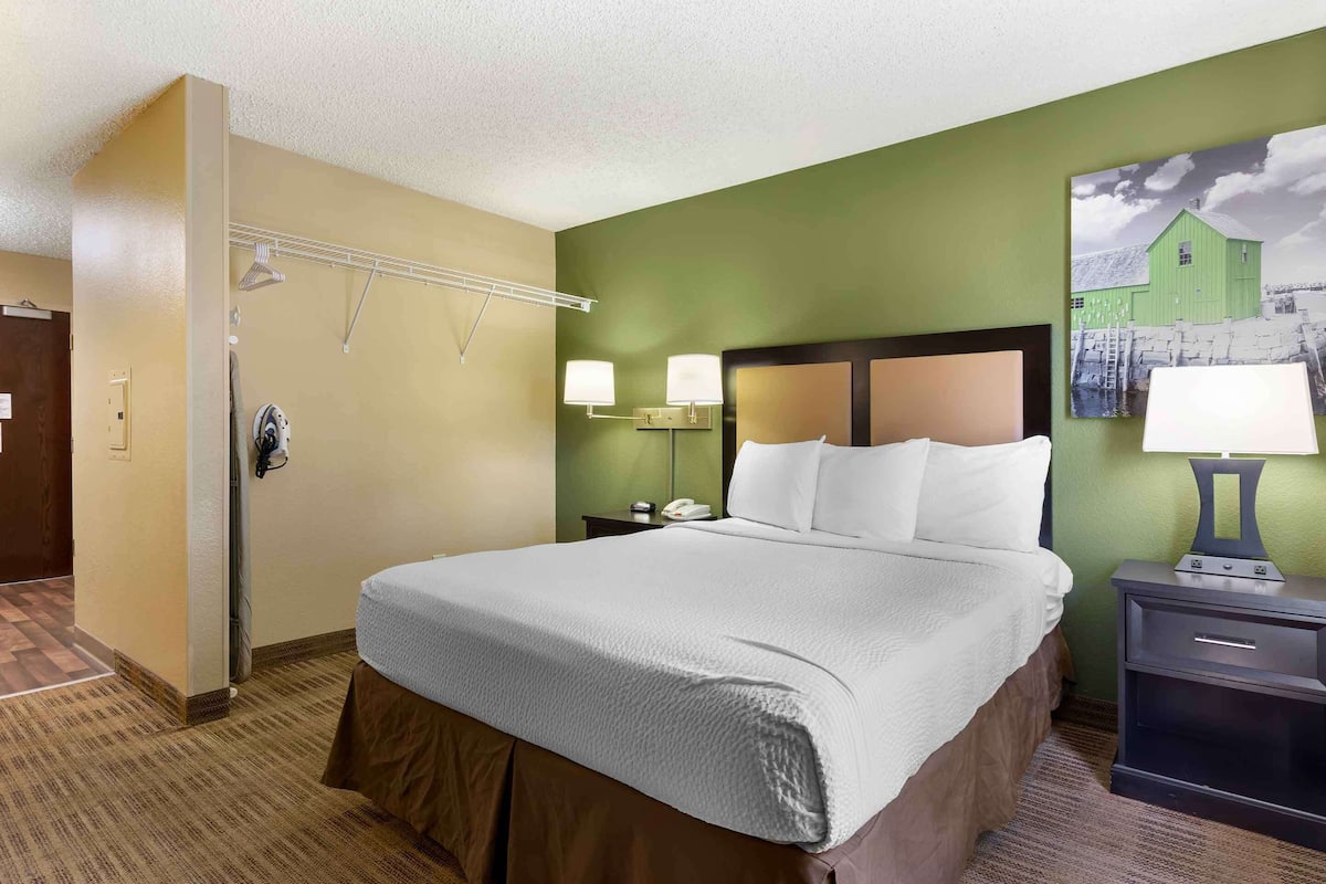 A hotel room with a bed, two bedside lamps, a nightstand, a wall-mounted coat rack, and a painting of a green barn.