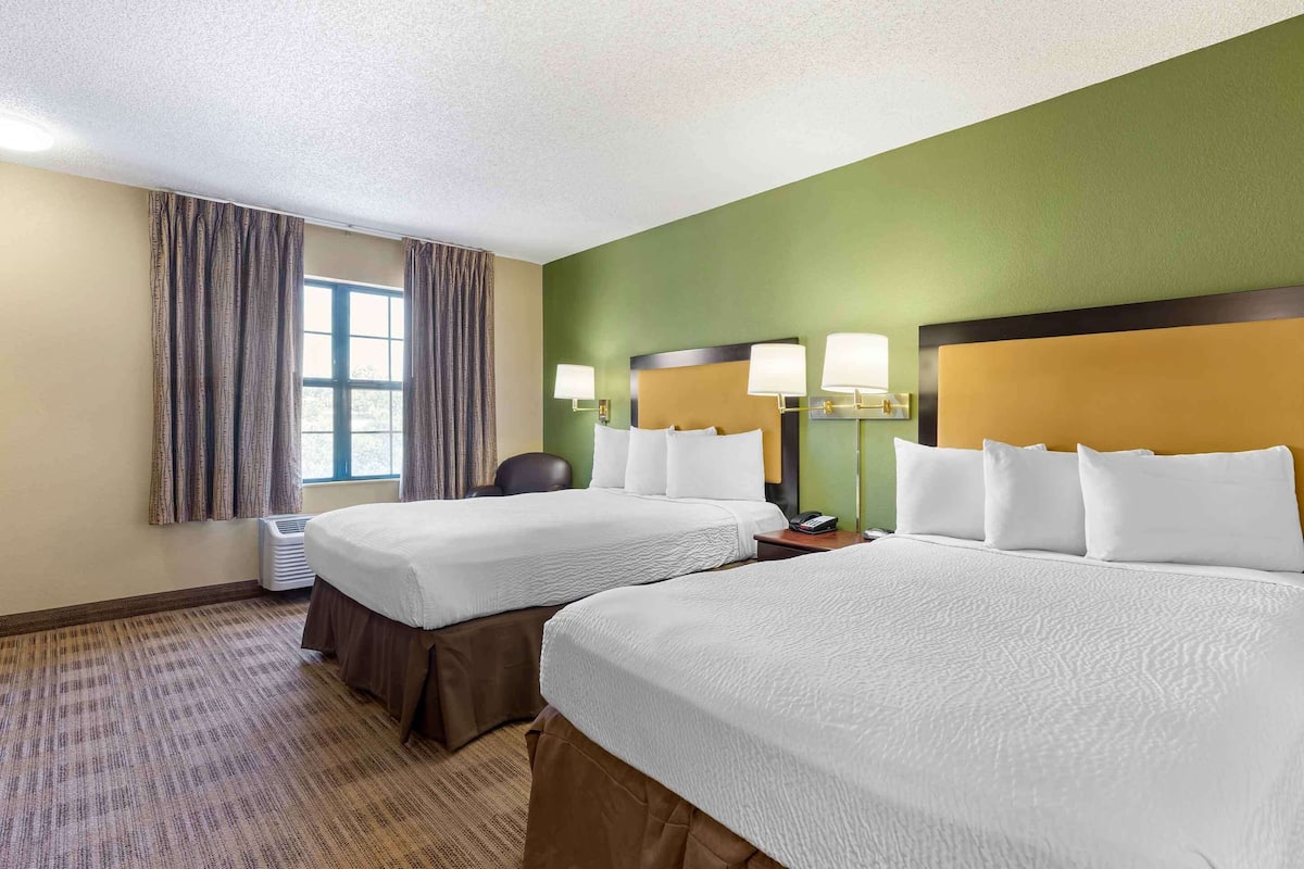 A hotel room with two beds, a window with curtains, a desk with a lamp, and a wall-mounted air conditioner.