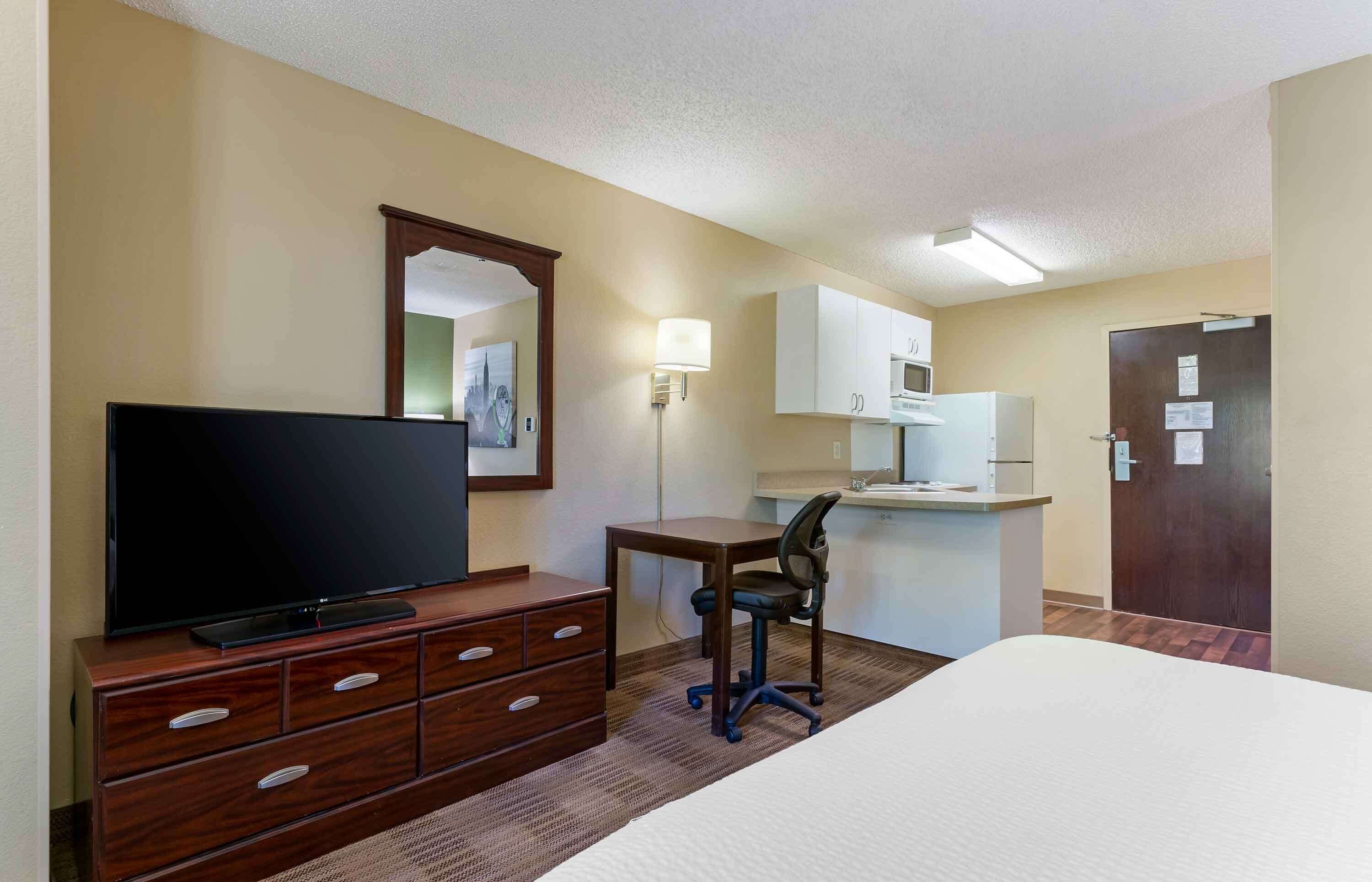 A hotel room with a bed, a TV on a wooden dresser, a desk with a chair, a microwave, and a door with a sign.