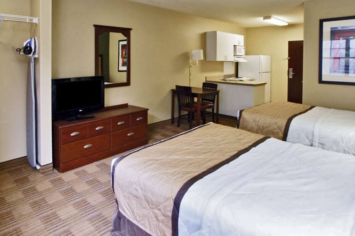 A hotel room with two beds, a desk, a chair, a television, and a kitchenette.