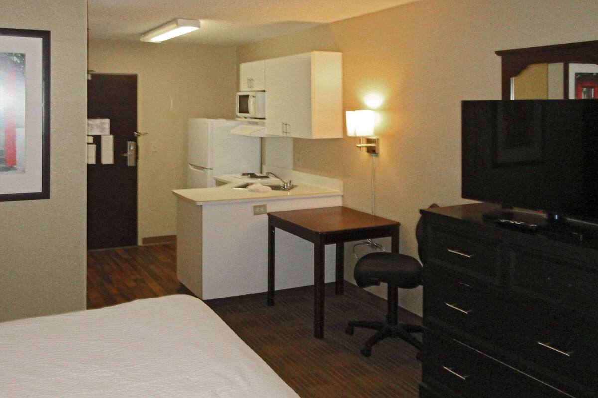 A hotel room with a bed, a small kitchenette, a desk, a chair, and a TV.