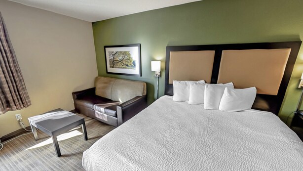 A hotel room with a large bed, a sofa, a small table, and a painting on the wall. - Suburban Studios Minneapolis Bloomington (Bloomington)