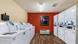 A laundry facility with multiple washing machines and dryers, a wall-mounted television, and a framed mirror. - Suburban Studios Minneapolis Bloomington (Bloomington)