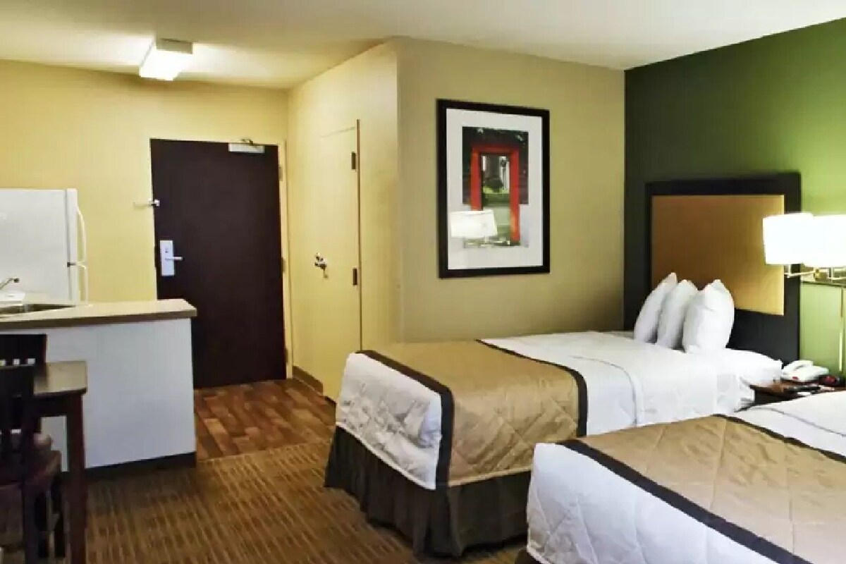A hotel room with two beds, a desk, a chair, a door, and a framed picture on the wall.