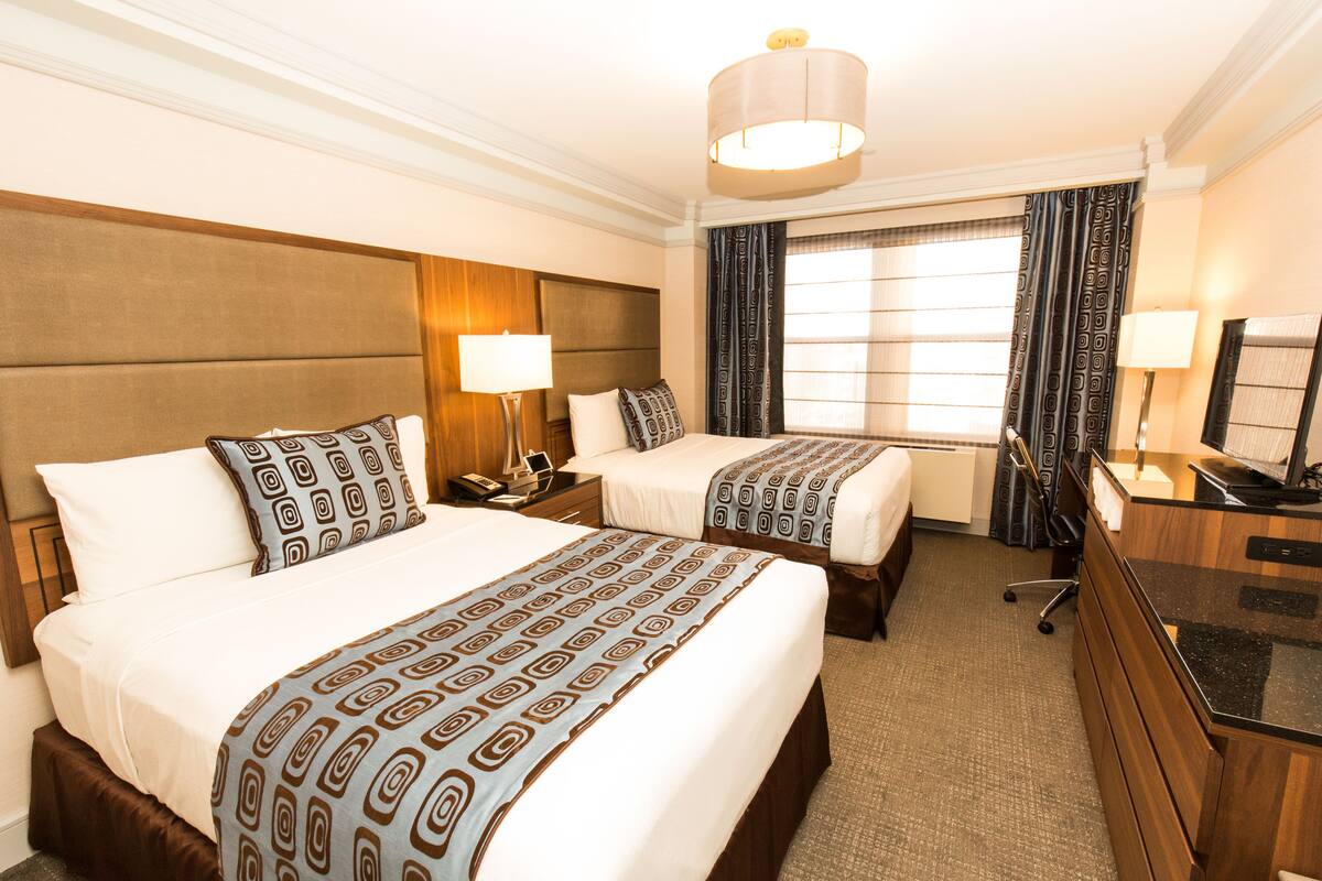 Quarto de hotel com duas camas, uma escrivaninha, uma cadeira, um abajur e uma janela grande com cortinas.