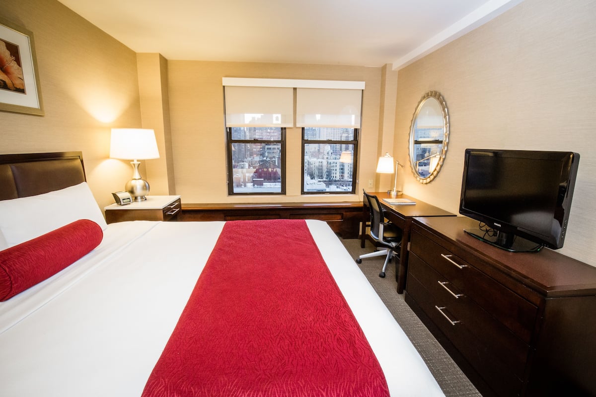 Quarto de hotel com cama, escrivaninha, televisão e janela com vista para a cidade.